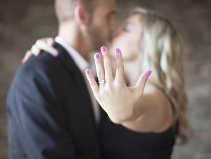 Woman’s hand featuring diamond engagement ring with couple kissing in background.