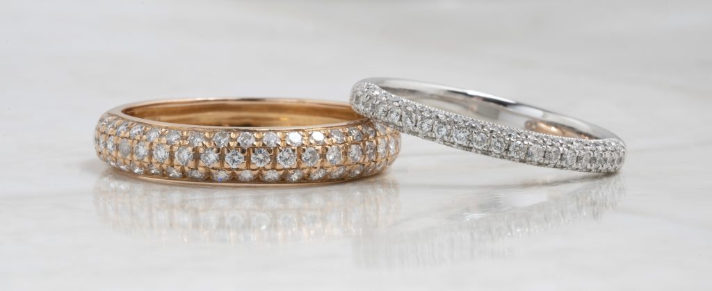 A rose gold and a white gold wedding band, both set with diamonds, stacked on a white table.