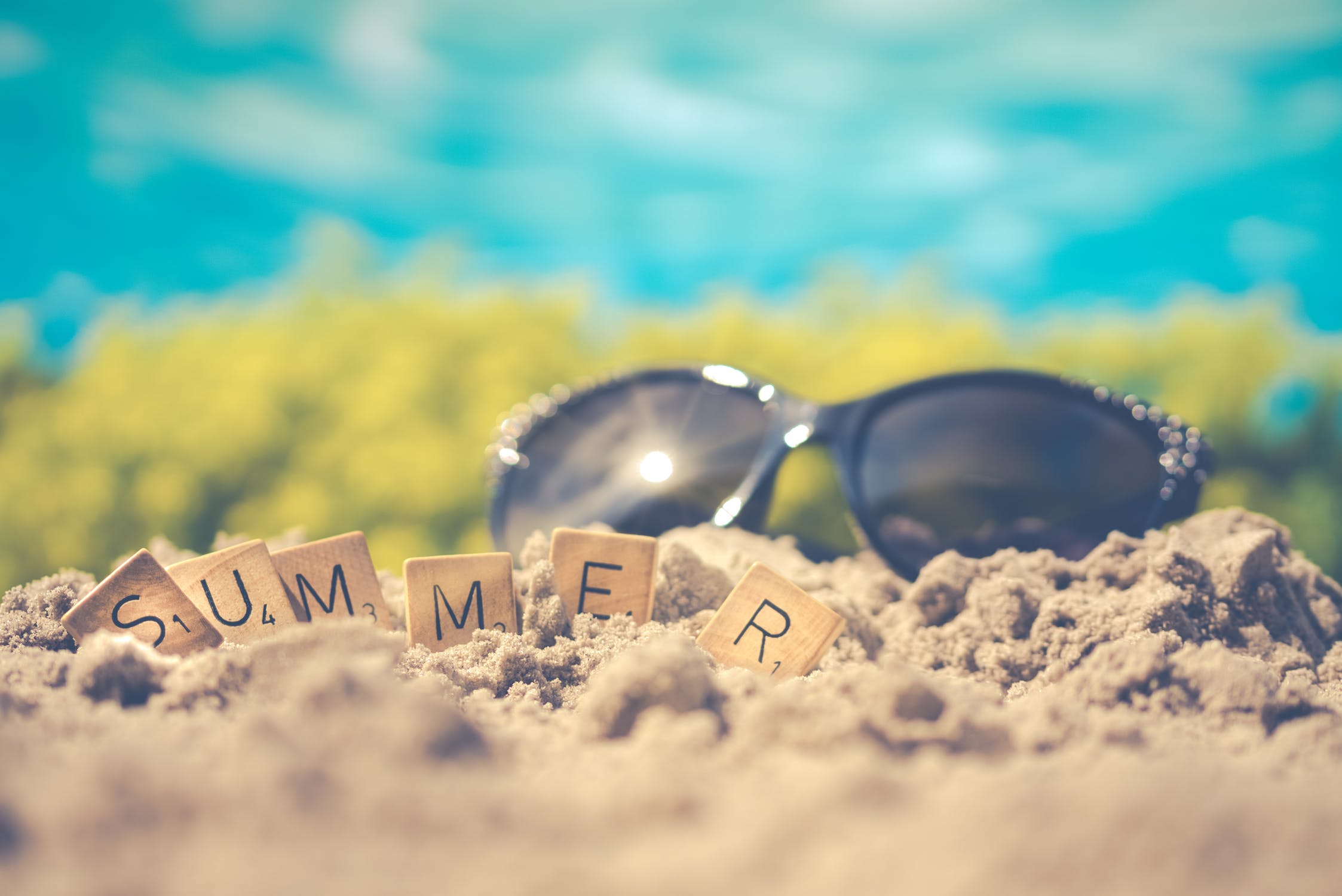 Black sunglasses in the sand at the beach with Scrabble tiles spelling out “Summer”.