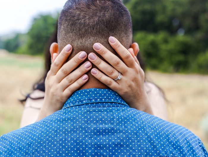 Woman’s hands on back of man’s head featuring her diamond engagement ring.