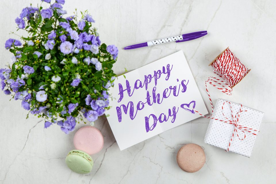 Mother’s Day card with two gift boxes and a vase of purple flowers on white table.