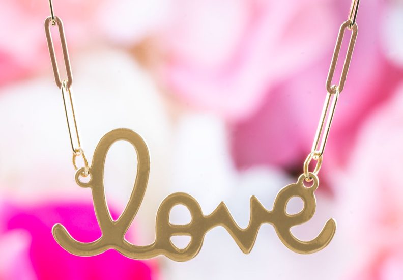 Yellow gold “LOVE” script necklace with elongated oval link chain on flower background.