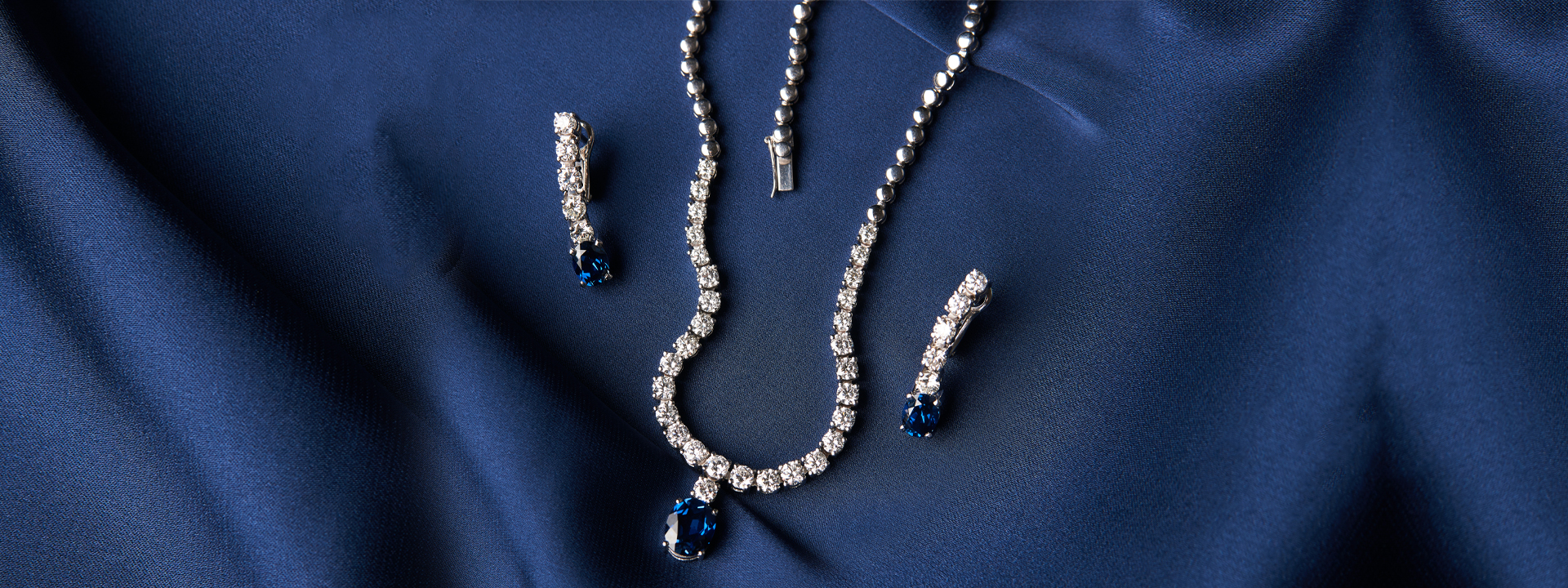 White gold necklace and earrings both set with diamonds and blue sapphires on a blue fabric background.