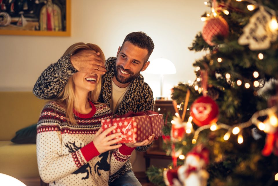 Man covering woman's eyes and giving her a gift in front of Christmas tree.