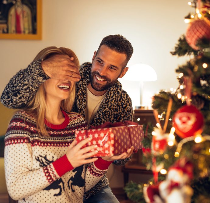 Man covering woman's eyes and giving her a gift in front of Christmas tree.