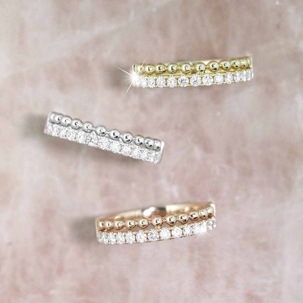 Three two-row diamond eternity bands in yellow gold, rose gold, and white gold on a tan
table.