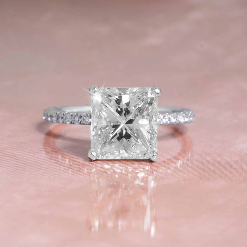 Princess-cut diamond engagement ring with a pavé band set in white gold, elegantly displayed on a soft pink background.