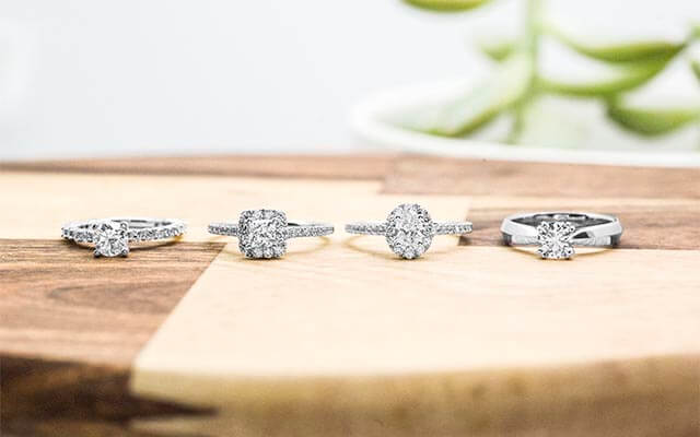 Four white gold diamond engagement rings of various shapes lined up on a wooden table.