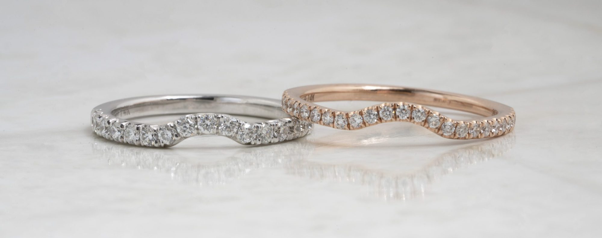 Two curved diamond wedding bands in white gold and rose gold on a white table.