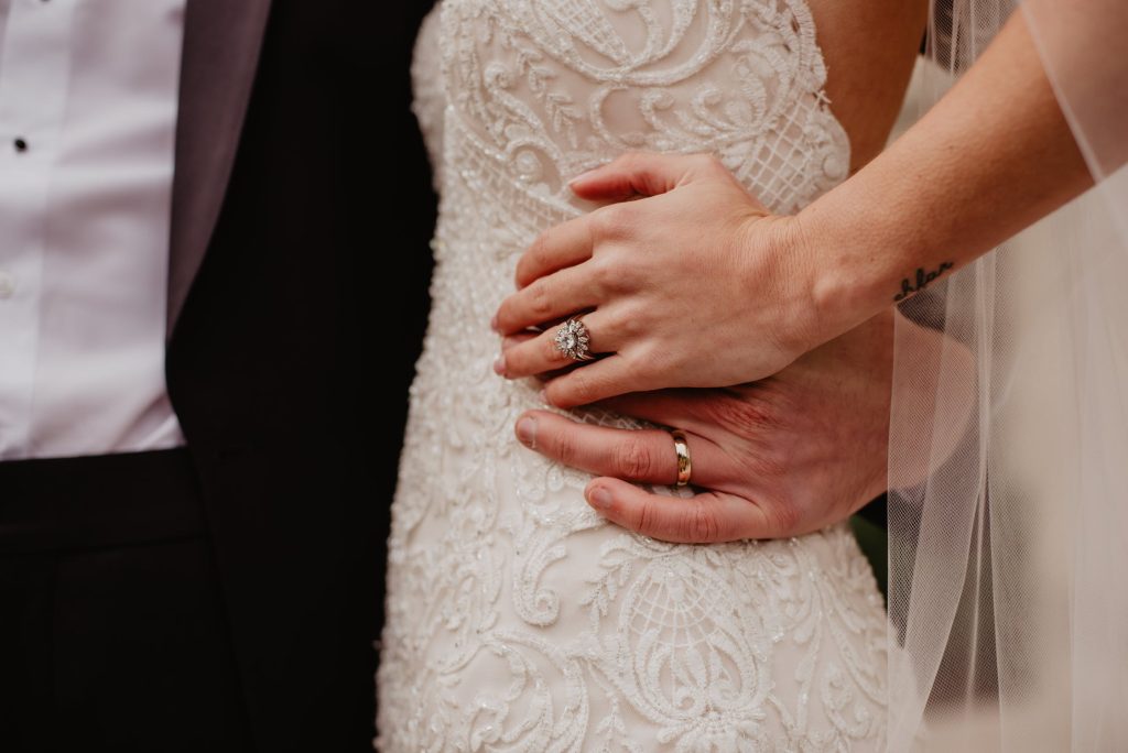 Bride and groom’s hands on bride’s waist featuring her diamond engagement ring and
their matching wedding bands.