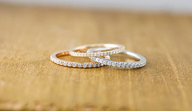 Three women’s eternity wedding bands in rose, yellow, and white gold set with diamonds
on a wooden table.